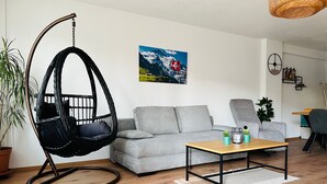 Ein modernes Wohnzimmer mit einem grauen Sofa, einem hängenden schwarzen Sessel, einem Couchtisch aus Holz und einem Gemälde von einer Berglandschaft.