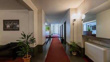Un couloir d’hôtel avec un tapis rouge, des plantes en pot, un canapé noir et un miroir fixé au mur.