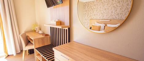 A modern hotel room with a wooden desk, a chair, a TV mounted on the wall, and a large mirror reflecting the bed.