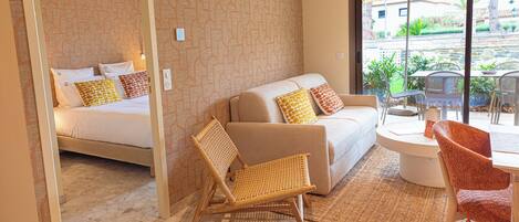 A hotel room with a bed, a sofa, a wicker chair, a dining table with chairs, and a view of the outdoors.