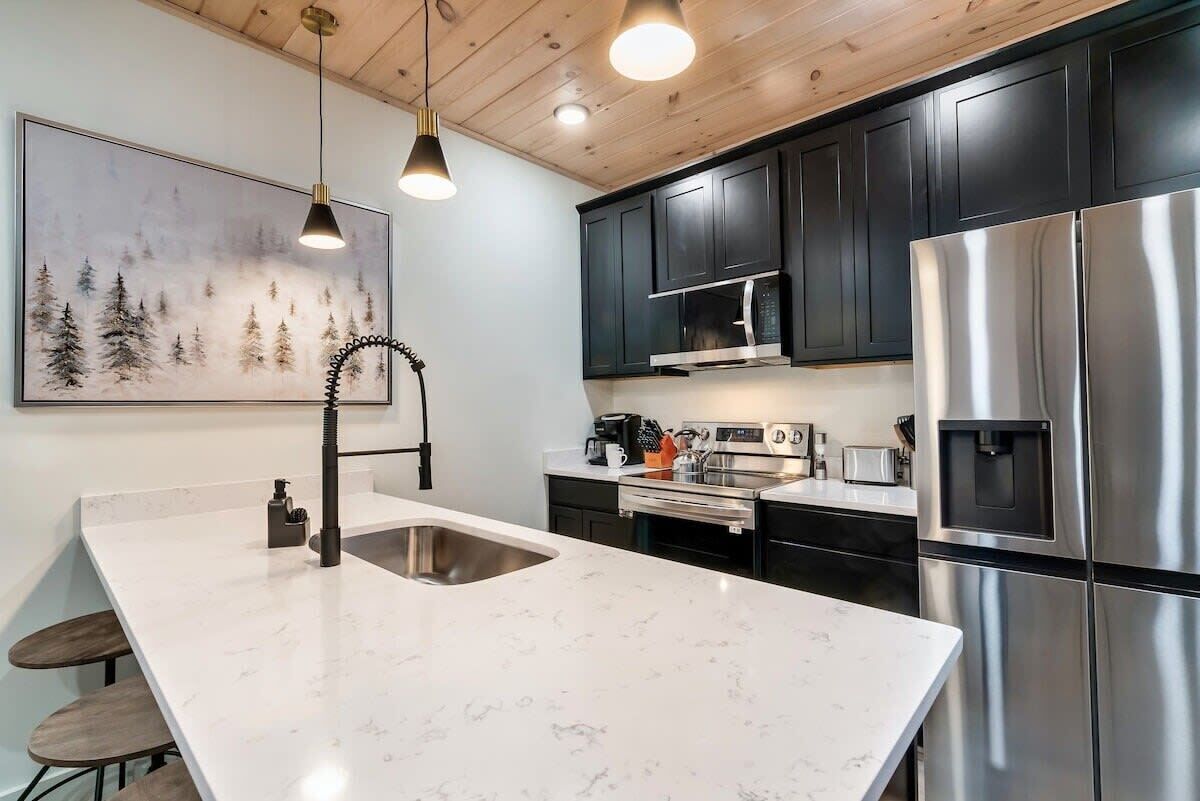 A modern kitchen with dark cabinets, stainless steel appliances, a marble countertop, and pendant lighting.