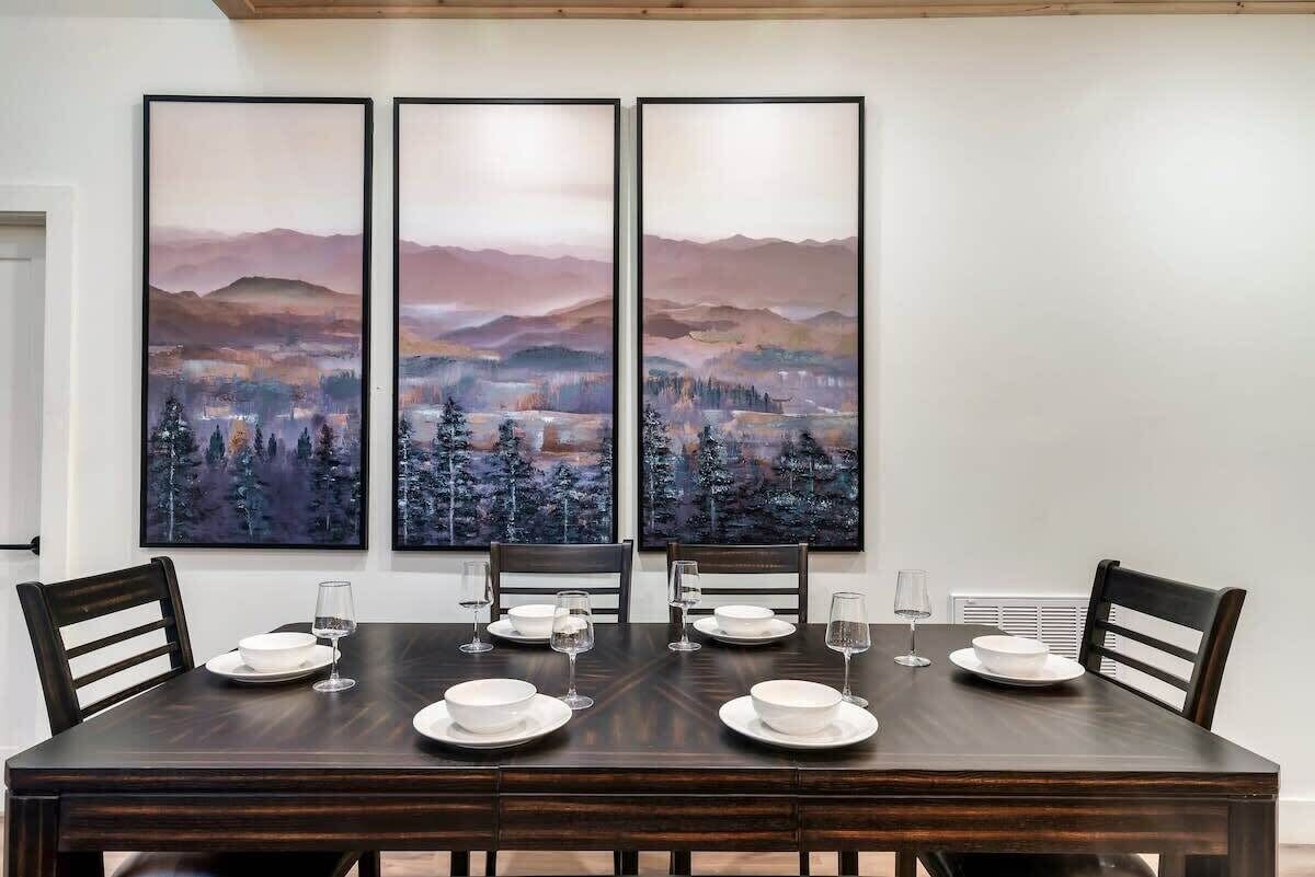 A dining room with a dark wooden table set for six, flanked by three large landscape paintings.