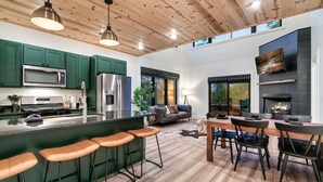 A modern kitchen with green cabinets, a large island with bar stools, a dining area with a fireplace, and a cozy seating area.