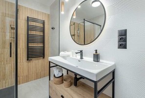 A modern bathroom with a white sink, black faucet, and a round mirror. There is a towel rack and a shower area visible.