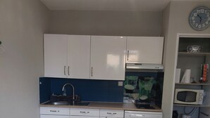 A compact kitchen with white cabinets, a sink, a microwave, and a clock on the wall. - Apartment terasse Le Barcarès  (Le Barcarès)