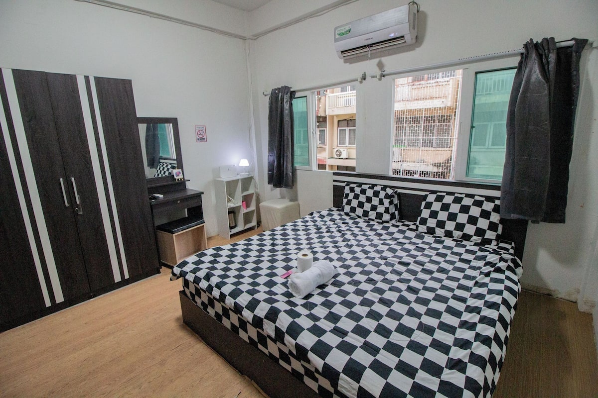 A bedroom with a checkered bedspread, a wardrobe, a desk with a lamp, and a window with curtains.