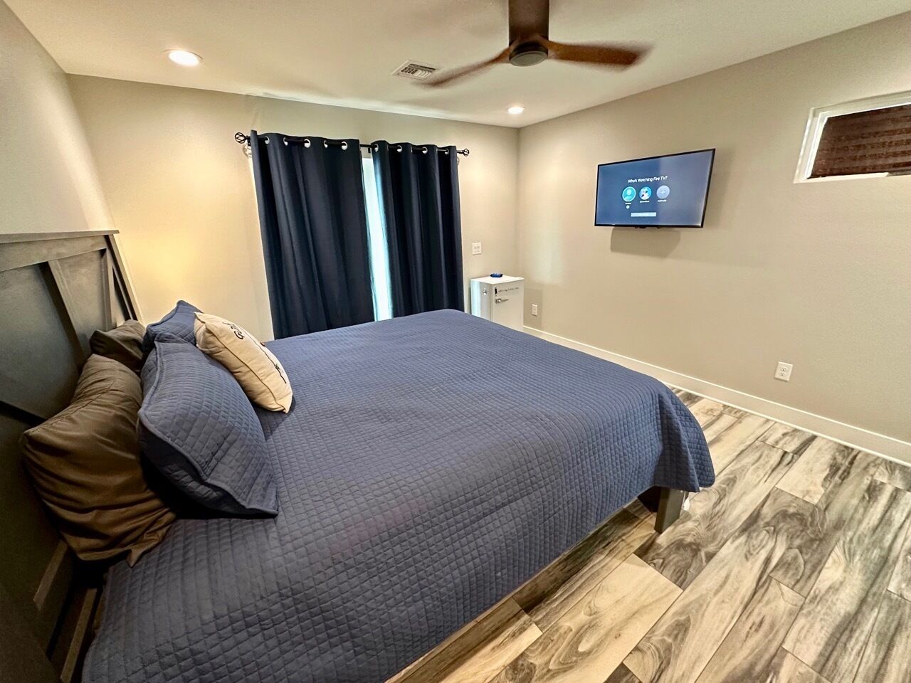 Une chambre à coucher avec un lit, des rideaux foncés, un ventilateur de plafond, une télévision et un plancher en bois.