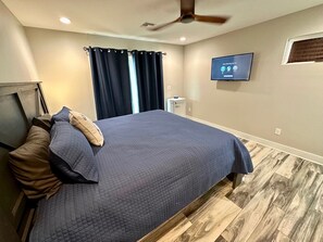 Camera da letto con un letto, tende scure, ventilatore a soffitto, televisore e pavimento in legno.