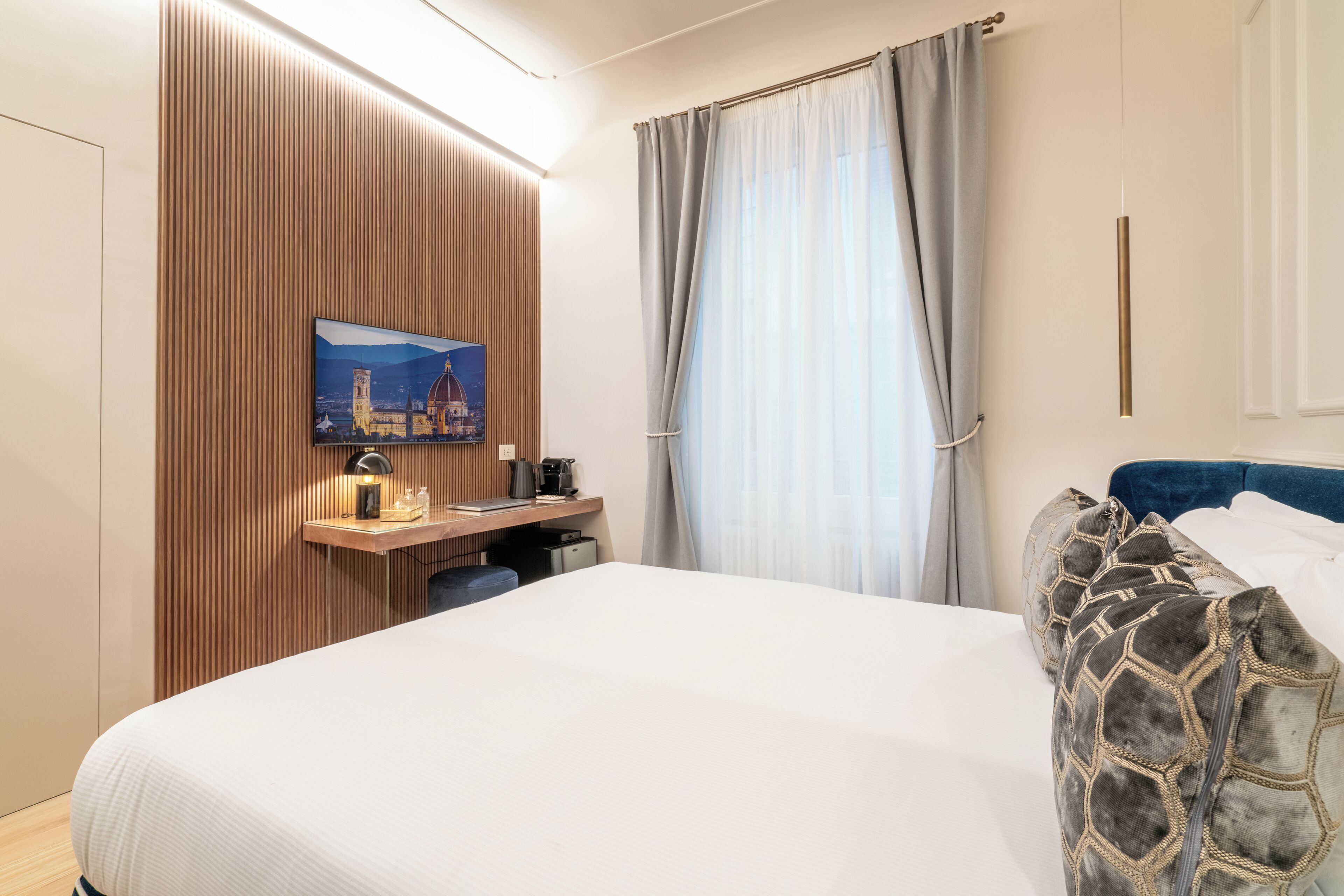 A hotel room with a bed, a desk with a lamp, a painting, and a window with curtains.