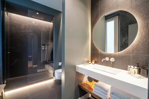 Une salle de bain moderne avec une douche à parois de verre, un lavabo blanc avec un miroir rond et un banc en bois sur lequel sont posées des serviettes.
