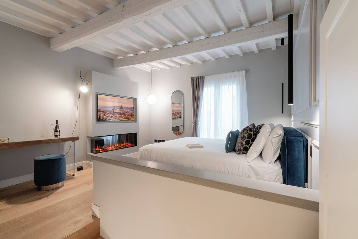 A modern bedroom with a bed, a fireplace, a cityscape painting, and a large window with curtains.