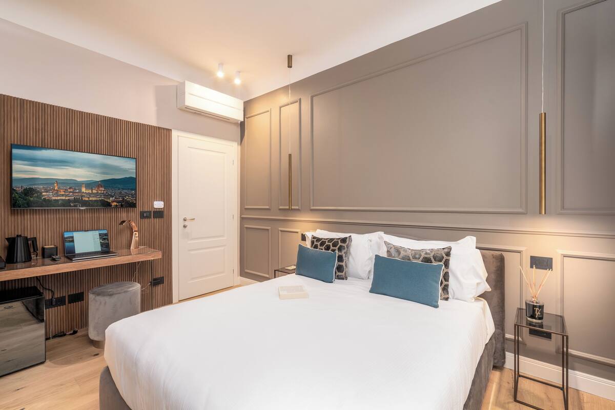 A modern hotel room with a large bed, a desk with a laptop, a TV mounted on the wall, and a small side table with a candle.