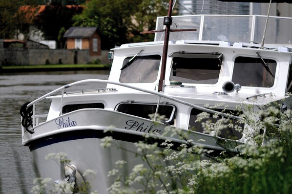 Un bateau nommé «
Phile
» accosta sur une rivière paisible, bordée d’un mur de pierres et d’une végétation luxuriante en arrière-plan.
