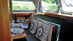A compact dining area with a wooden table set for two, featuring plates, glasses, and a bottle. Two cushions with nautical designs are placed on the bench.