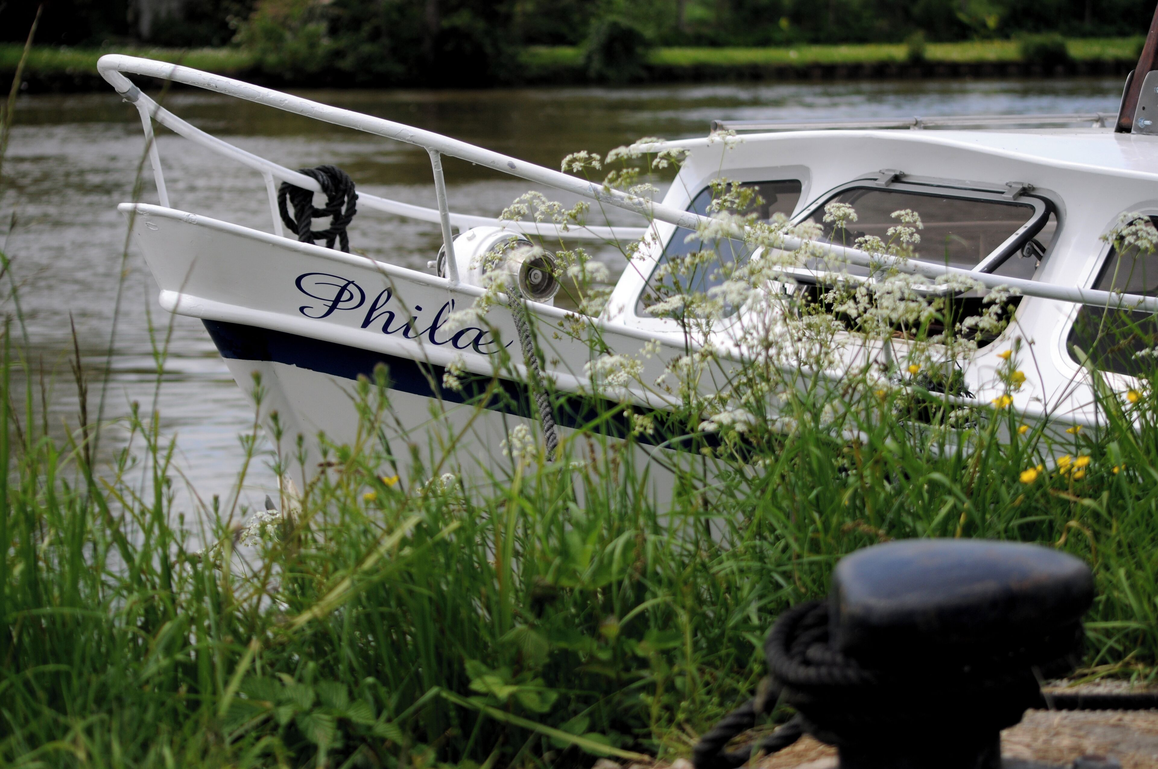 A boat named 'Phila' docked at a calm river with lush greenery around.