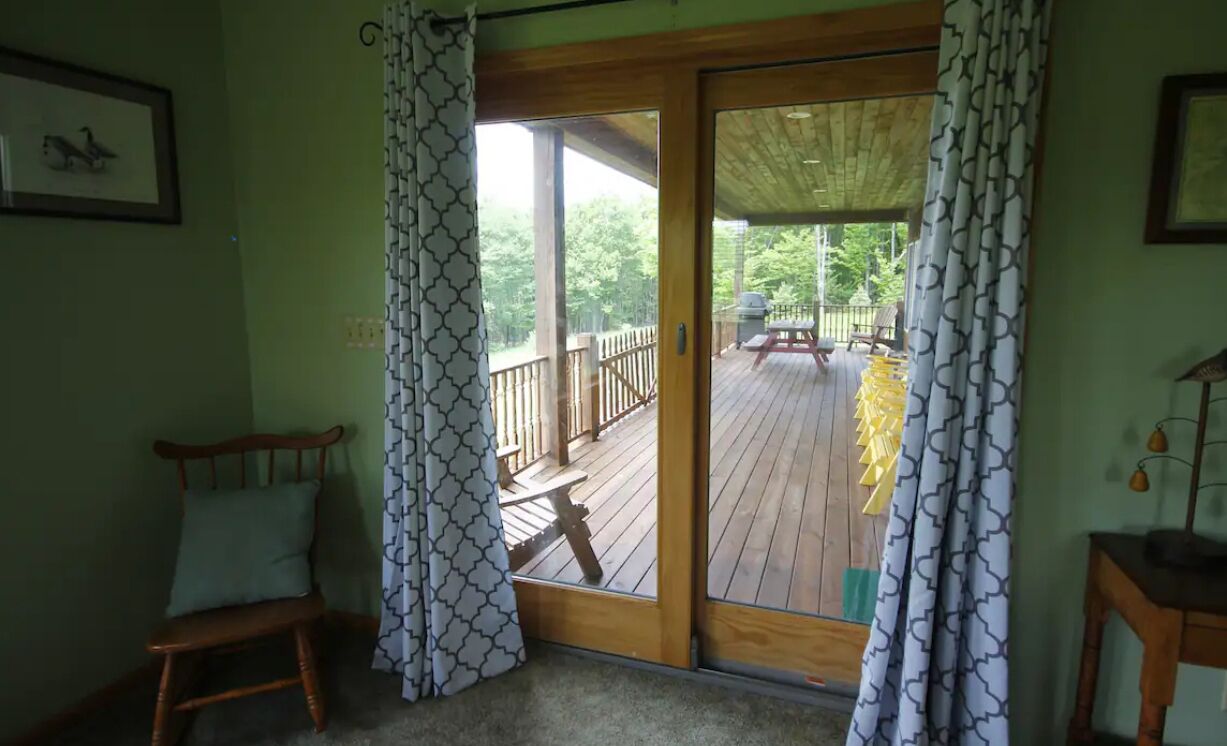 Ein Zimmer mit Holzstuhl, einer Bank und einem Holztisch. Eine Glastür führt zu einer Terrasse mit Picknicktisch und Grill.