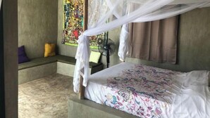 A bedroom with a canopy bed, a small bedside table, a fan, and a colorful artwork on the wall. - Bien Bassile 3 (Guéréo)
