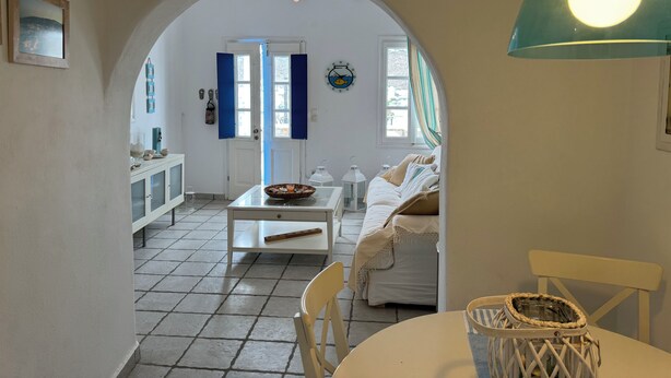 Un interior acogedor con una zona de comedor, una cocina visible a través de un arco, y una cama con cabecero blanco.