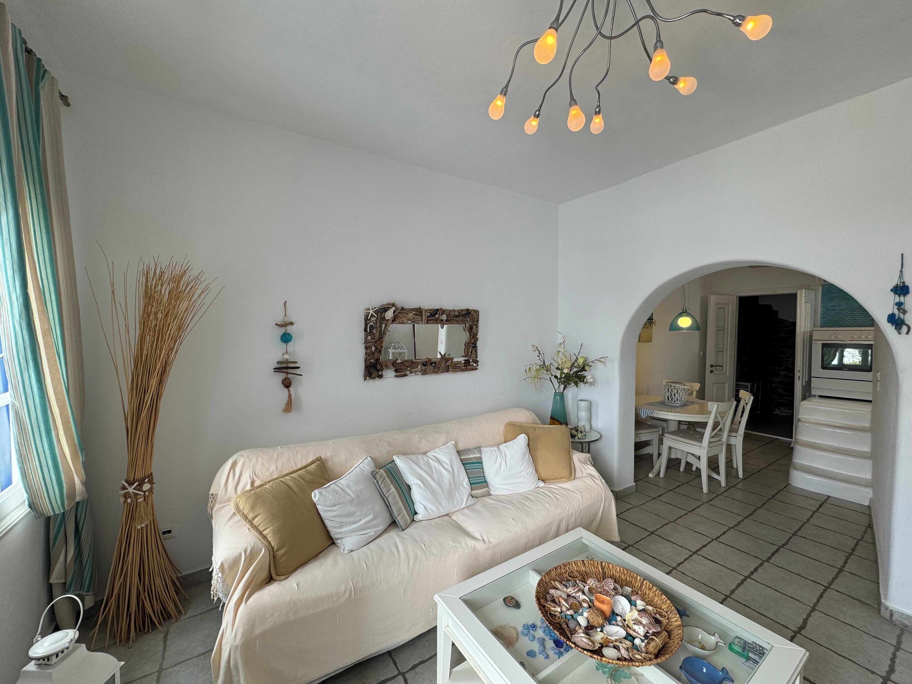 A living room with a sofa, a coffee table with decorative items, a hanging light fixture, and a dining area with chairs and a table.