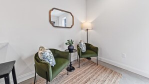 A room with two green armchairs, a small side table with a plant, a floor lamp, and a mirror on the wall.