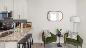 A modern kitchen with white cabinets, a granite countertop, stainless steel appliances, and two green armchairs with patterned cushions.
