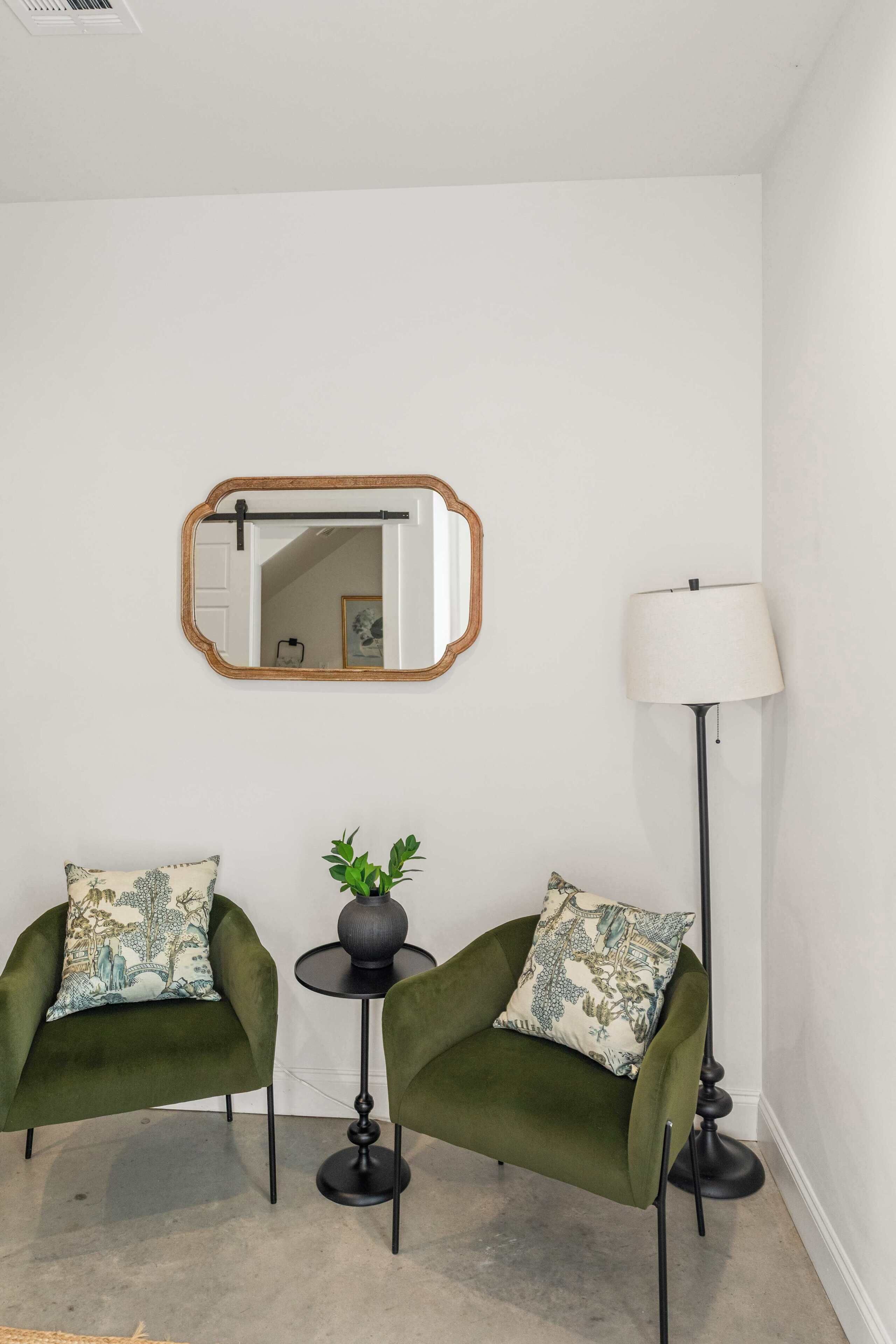 Una habitación con dos sillones verdes, una mesita redonda con una planta, una lámpara de pie y un espejo en la pared.