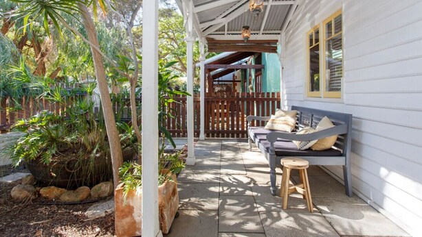 A covered porch with a bench and stool, surrounded by tropical plants.