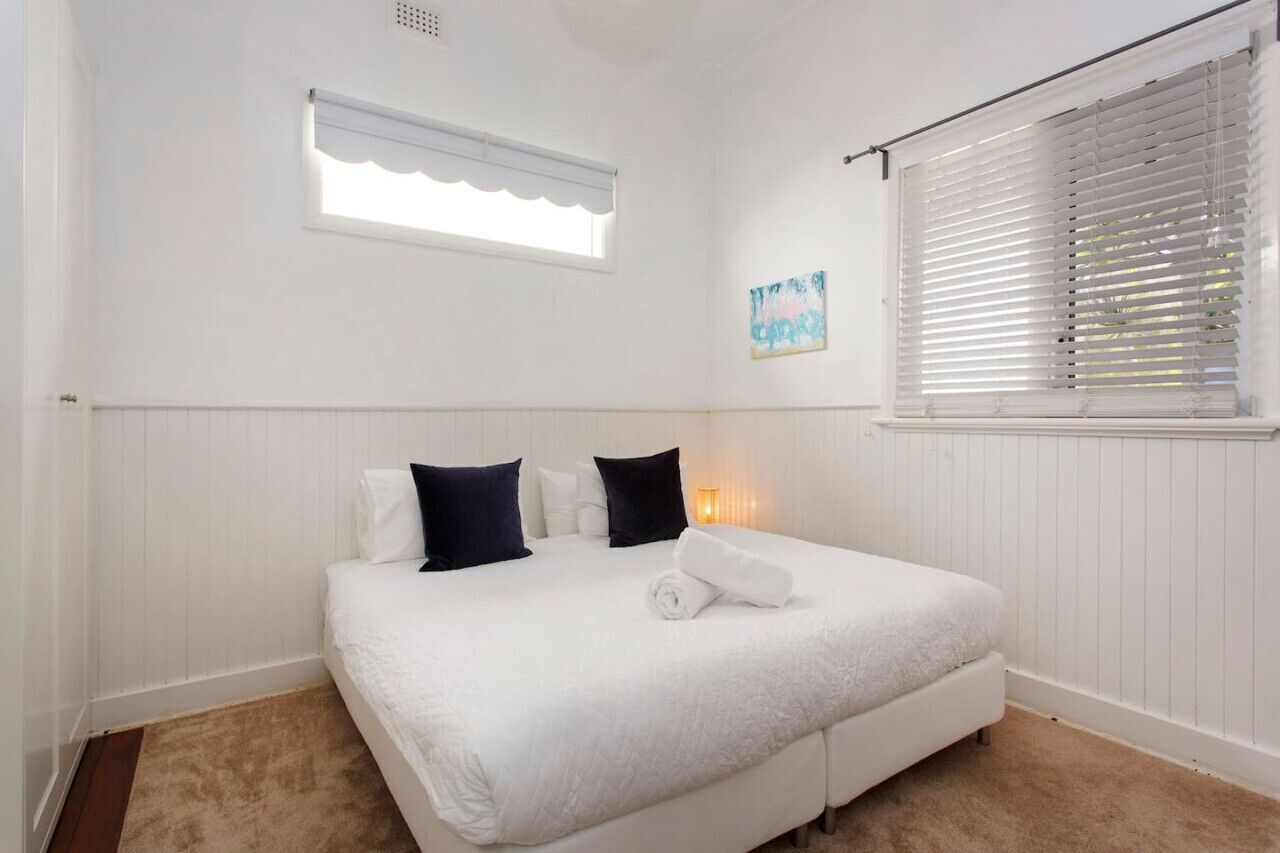 Une chambre à coucher avec un lit, des murs blancs, une fenêtre avec des stores, une petite fenêtre agrémentée d’une lambrequin blanche et un tableau accroché au mur.
