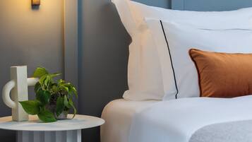 Een net opgemaakt bed met witte beddengoed en één bruin kussen, een klein witte nachtkastje met een plant, en een wandlamp.