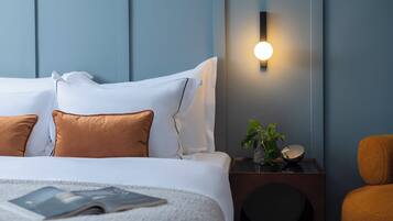 A neatly made bed with white and brown pillows, a bedside table with a plant and a bowl, and a wall-mounted light fixture.