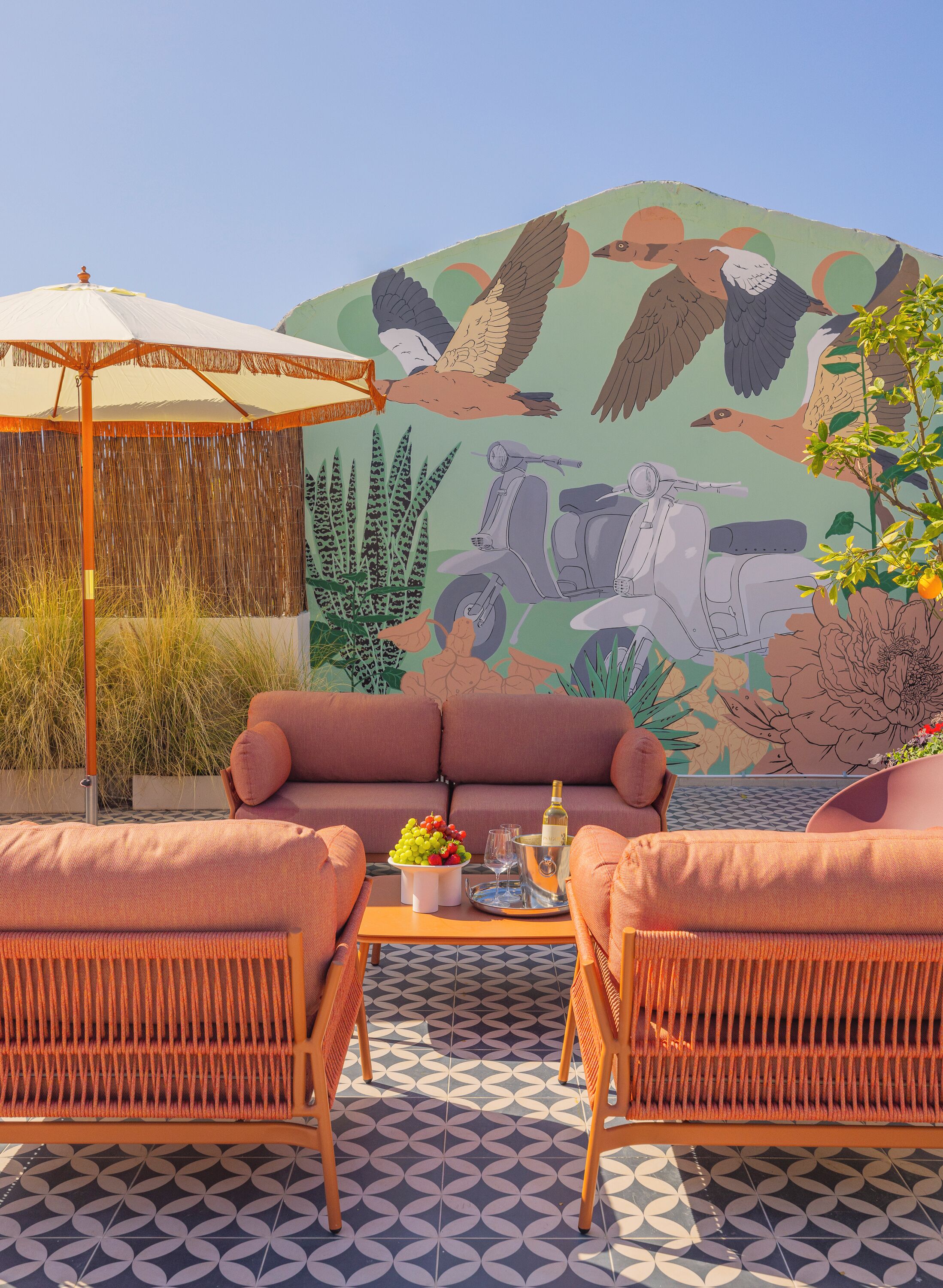 A cozy outdoor seating area with wicker furniture, a table with drinks and fruit, and a mural of birds and scooters in the background.