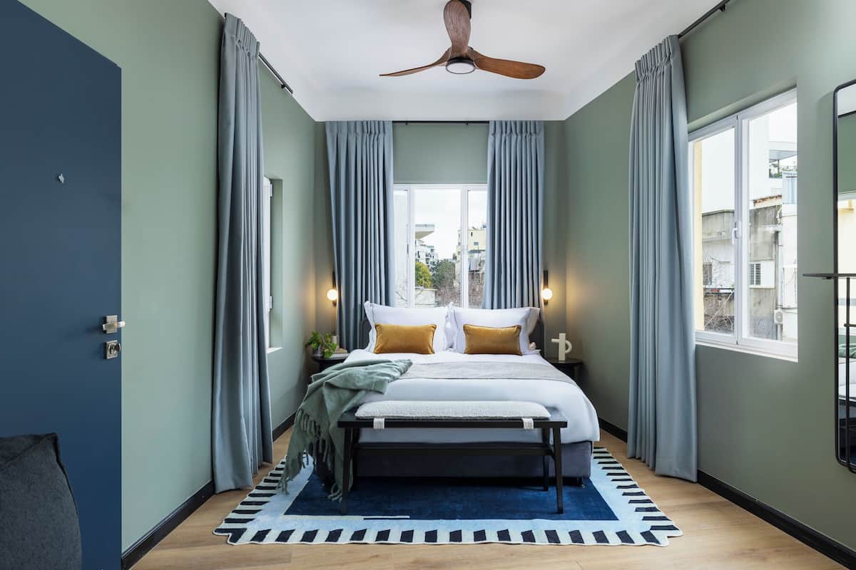 A bedroom with a bed, a ceiling fan, a window with curtains, and a view of buildings outside.