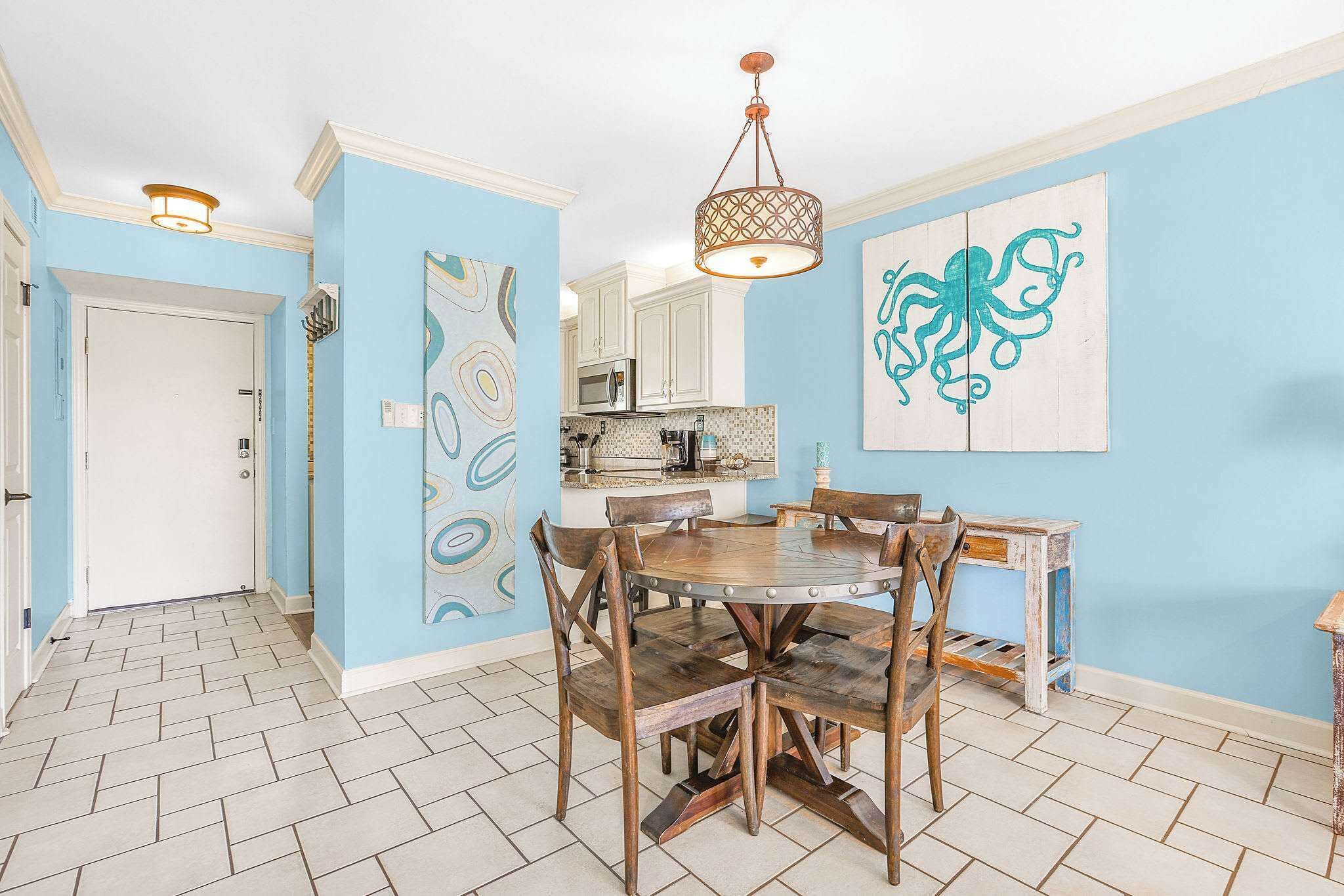 A dining area with a wooden table and chairs, a hanging light fixture, and a kitchen in the background.