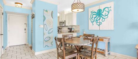 A dining area with a wooden table and chairs, a hanging light fixture, and a kitchen in the background.