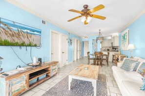 Sala de estar com ventilador de teto, televisão, mesa de centro, sofá e cozinha ao fundo.