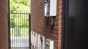 Une ruelle étroite bordée d’un mur de briques, avec une porte en métal et des boîtiers électriques fixés au mur.