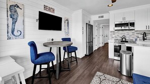 A modern kitchen with white cabinets, a stainless steel refrigerator, and a microwave. There is a dining area with blue chairs and a round table.