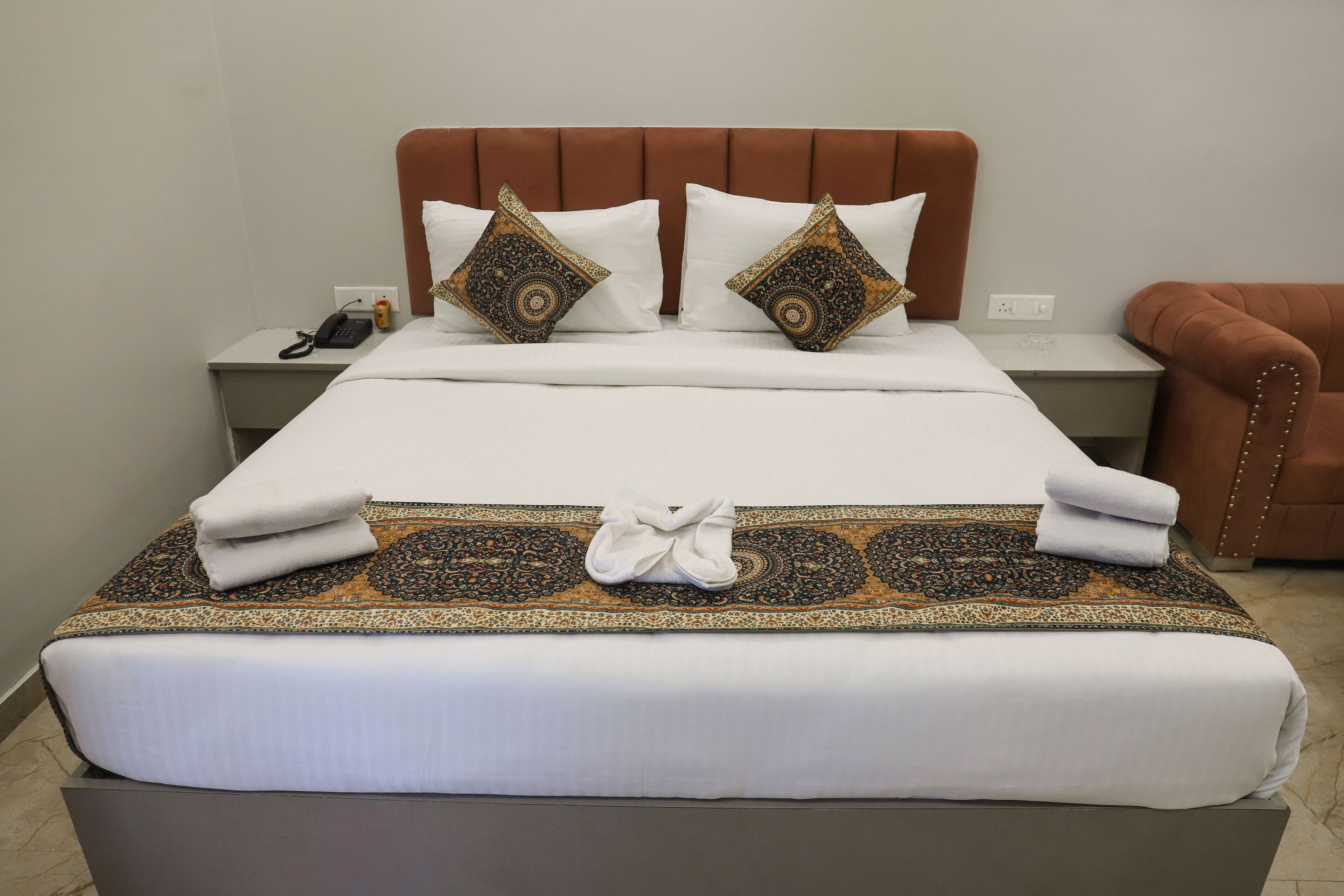 Una cama bien hecha con sábanas blancas y almohadones decorativos, una mesita de noche con un teléfono y una lámpara, y un sillón tapizado en marrón.