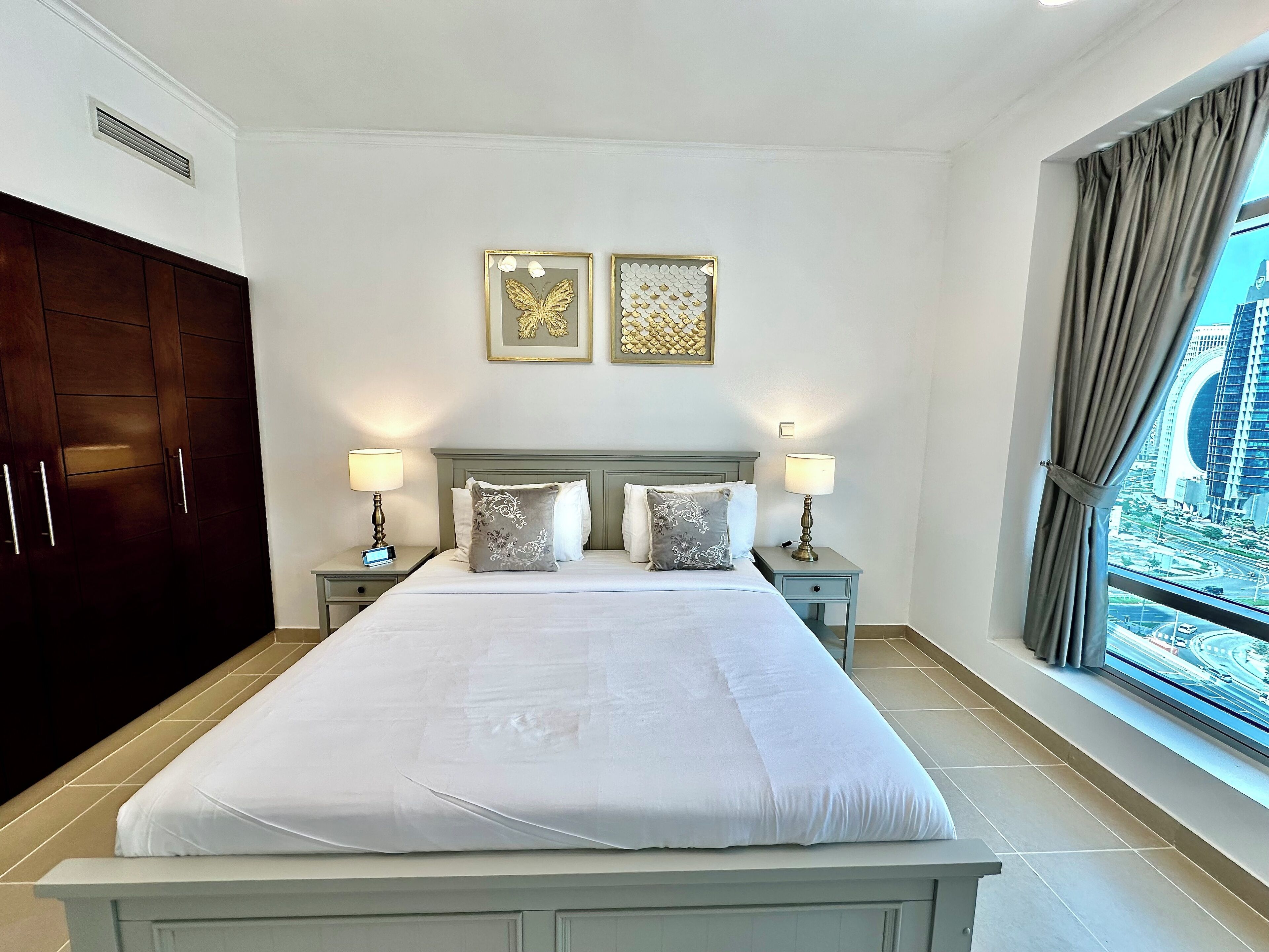 A hotel room with a large bed, two bedside tables with lamps, a wardrobe, and two framed pictures on the wall.