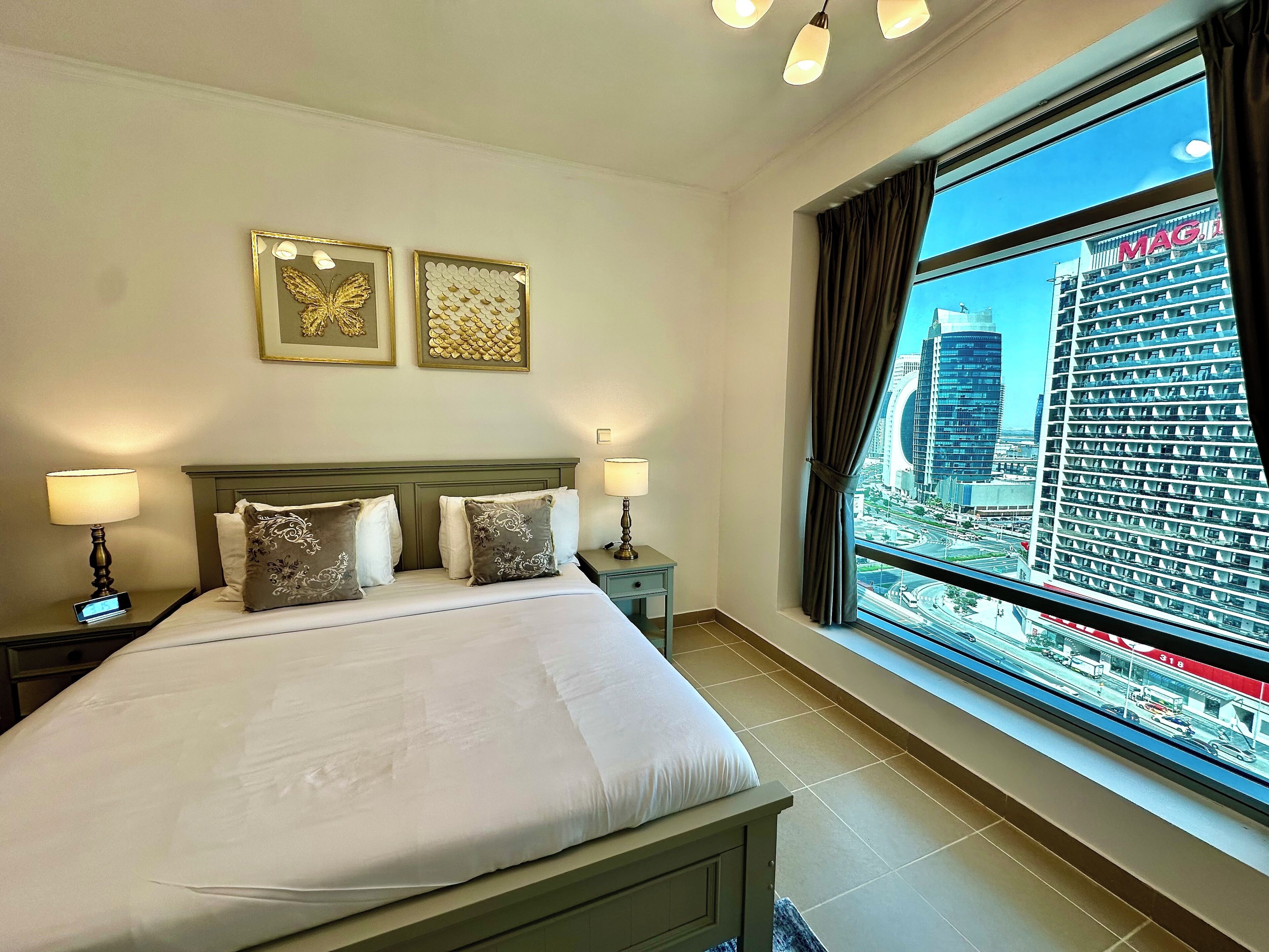 A hotel room with a large bed, two bedside tables with lamps, a view of a cityscape through a large window, and two framed artworks on the wall.