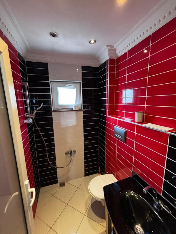 Un baño con paredes cubiertas de azulejos rojos y negros, un inodoro blanco, un lavabo negro y una ducha con cabezal de ducha manual.