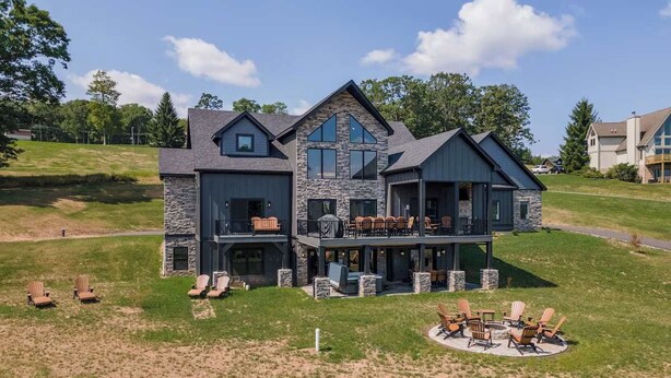 Une grande maison moderne avec une façade en pierre, plusieurs niveaux et un vaste patio extérieur aménagé avec du mobilier de jardin.