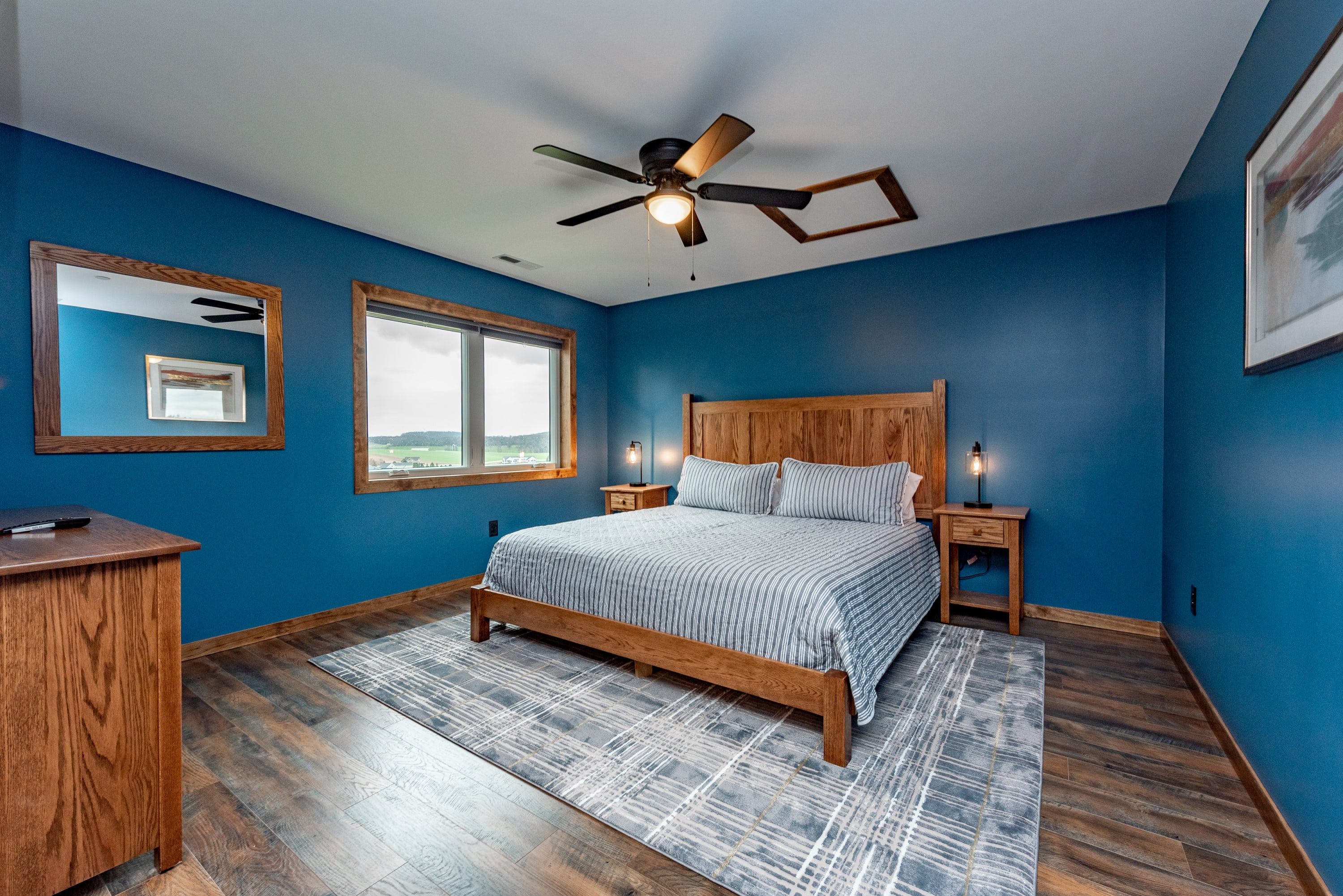 A bedroom with a bed, wooden headboard, nightstands, a desk, a ceiling fan, and a window with a view.