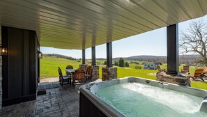 Un bain à remous installé sous une terrasse couverte, avec vue sur un champ verdoyant et des maisons au loin.
