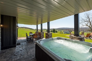 A hot tub under a covered patio with a view of a green field and houses in the distance. - The Crooked Oak Lodge - Deep Creek Lake - Indoor Pool & Hot Tub (Deep Creek, Maryland)