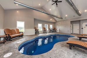 A swimming pool with tiled edges, wooden benches, and chairs around it. - The Crooked Oak Lodge - Deep Creek Lake - Indoor Pool & Hot Tub (Deep Creek, Maryland)