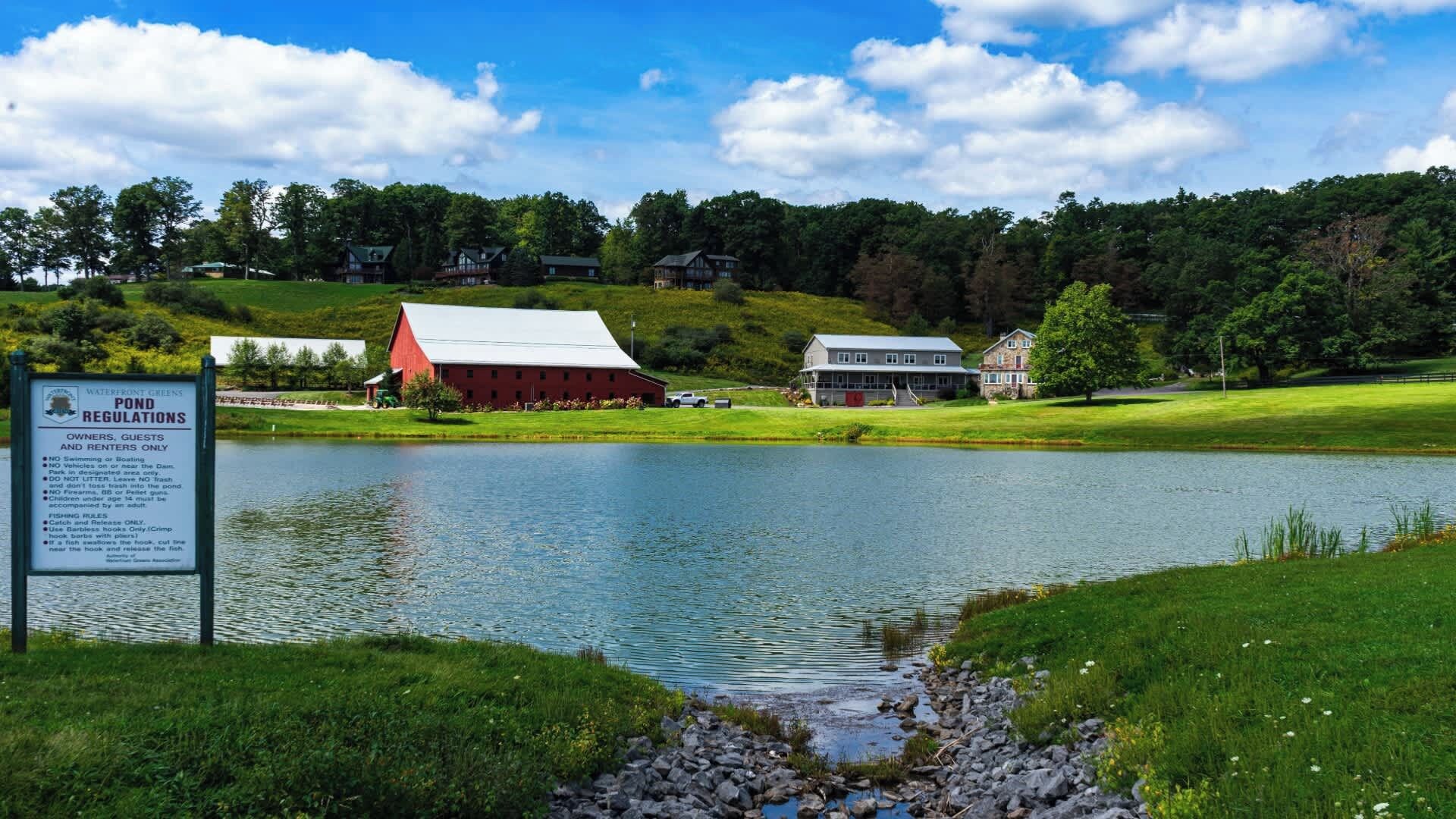 A pond with a sign detailing regulations, surrounded by a grassy area, a red barn, and a house.