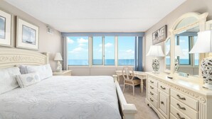 Une chambre avec un grand lit, un bureau et une commode. Les murs sont ornés de tableaux encadrés, il y a un miroir et, par les fenêtres, on peut admirer la vue sur l’océan.