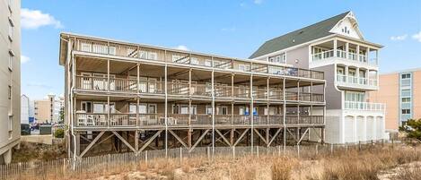 Un immeuble à plusieurs étages doté d’une terrasse en bois et de balcons, entouré d’herbe sèche et de sable.
