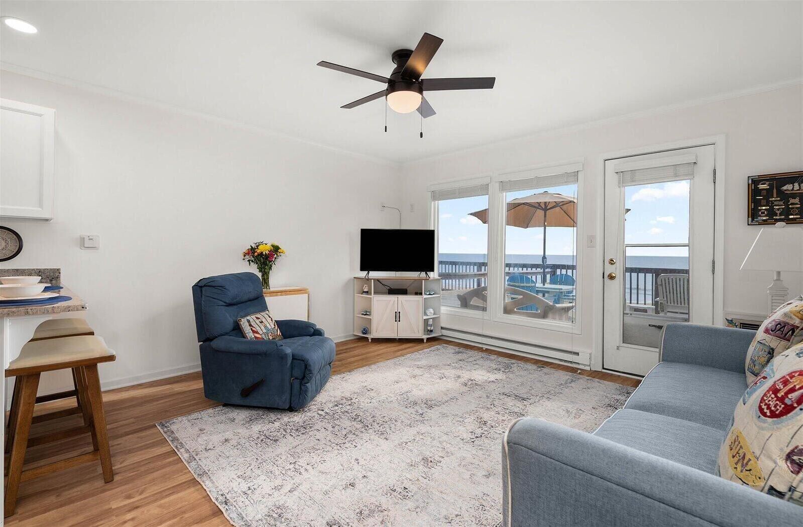 Un salon doté d’un ventilateur de plafond, d’un téléviseur, d’un coin repas avec une table et des chaises, et offrant une vue sur l’océan grâce à de grandes fenêtres.